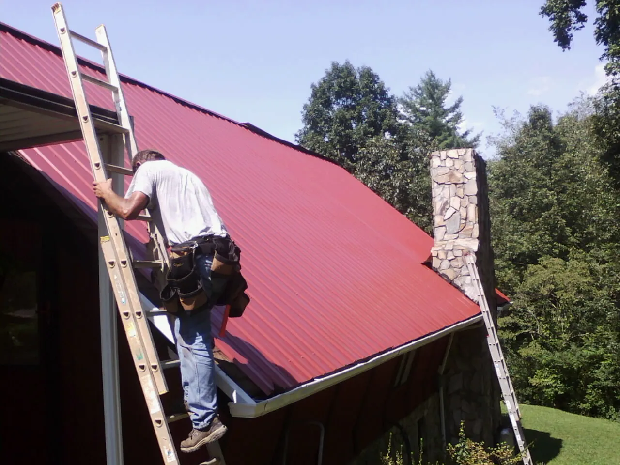 Roofer on a ladder beside a red metal roof and stone chimney — Soffit & Fascia Repair by Roofing Brothers in Smithton